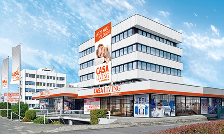 Das Bild zeigt das Gebäude von „Casa Living by Schaffrath“ in einem modernen, weißen Design mit orangefarbenen Akzenten. Das Gebäude hat mehrere Stockwerke und eine klare, kubische Architektur. Im oberen Bereich ist ein großes Banner mit der Aufschrift „Jetzt neu erleben“ sowie einem Bild einer lächelnden Frau und dem „Casa Living“-Logo angebracht. Im Erdgeschoss befinden sich große Schaufenster, durch die Möbel und Wohnaccessoires zu sehen sind. Links im Bild stehen mehrere weiße Fahnenmasten mit dem „Casa Living“-Logo. Vor dem Gebäude befinden sich ein gepflasterter Weg und gepflegte Grünflächen. Der Himmel ist blau mit dekorativen Wolken und vermittelt eine helle, freundliche Atmosphäre.